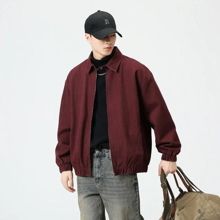 Man wearing burgundy bomber jacket, black cap, black shirt, gray jeans, holding brown duffel bag