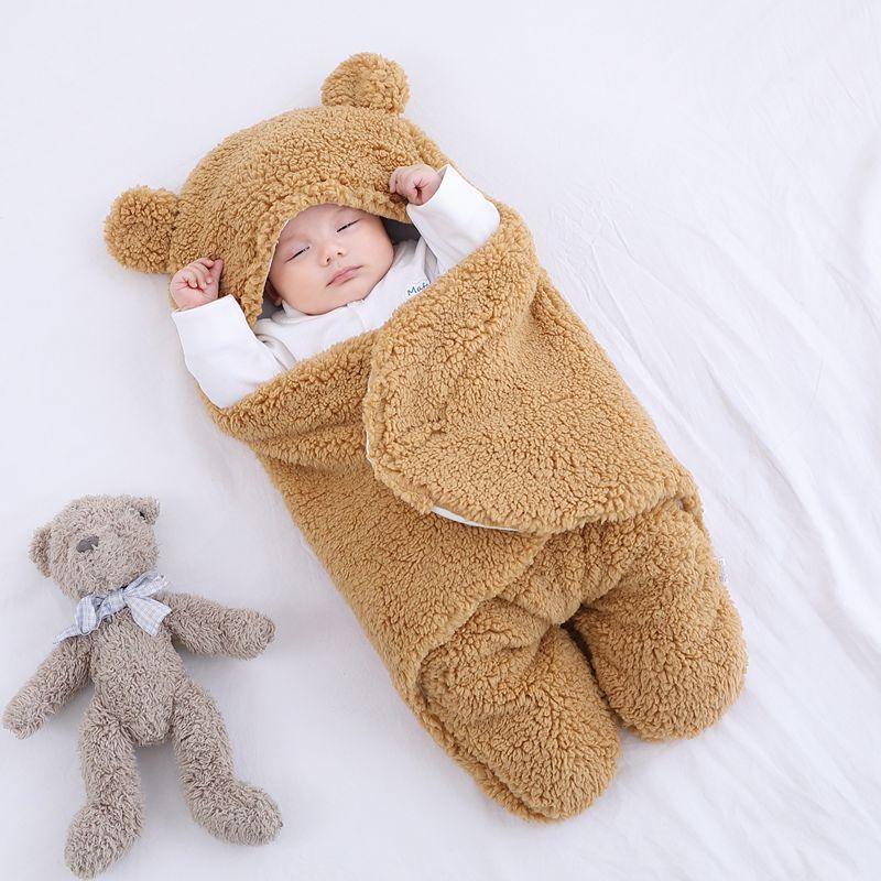 Sleeping baby wrapped in soft brown teddy bear fleece blanket with hood on white bed next to gray teddy bear