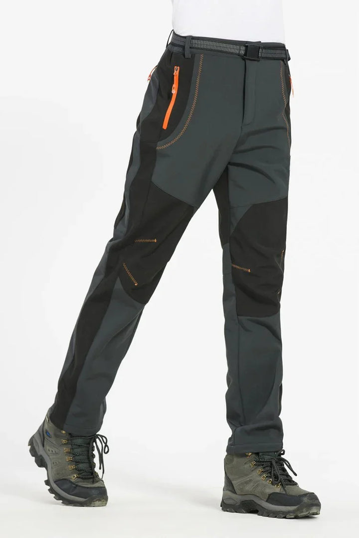 Men's hiking pants in dark gray with black panels and orange zippers, paired with gray hiking boots