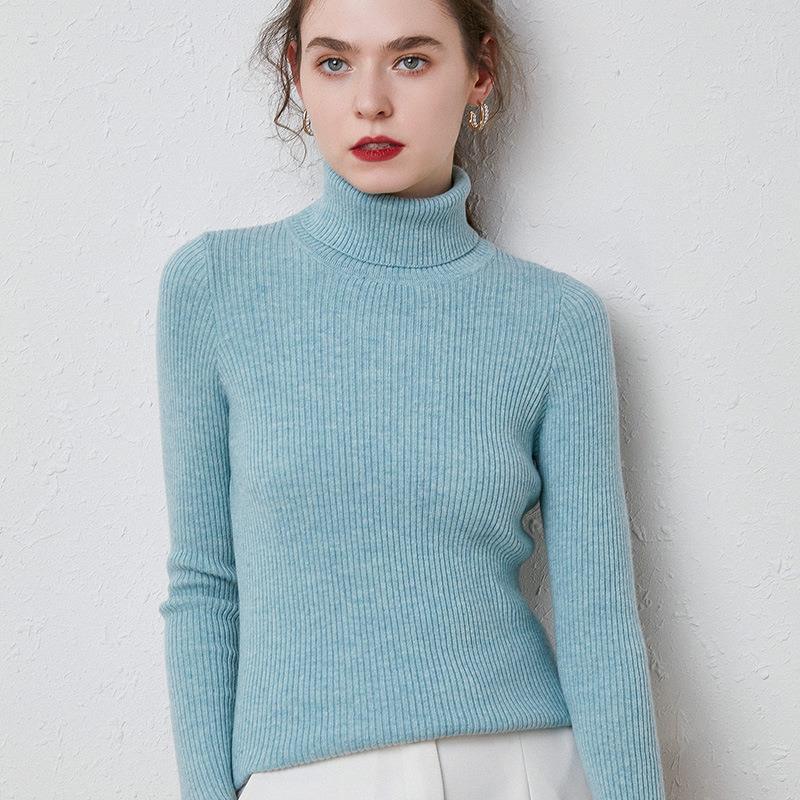 Woman wearing a light blue ribbed turtleneck sweater with long sleeves standing against a white textured wall