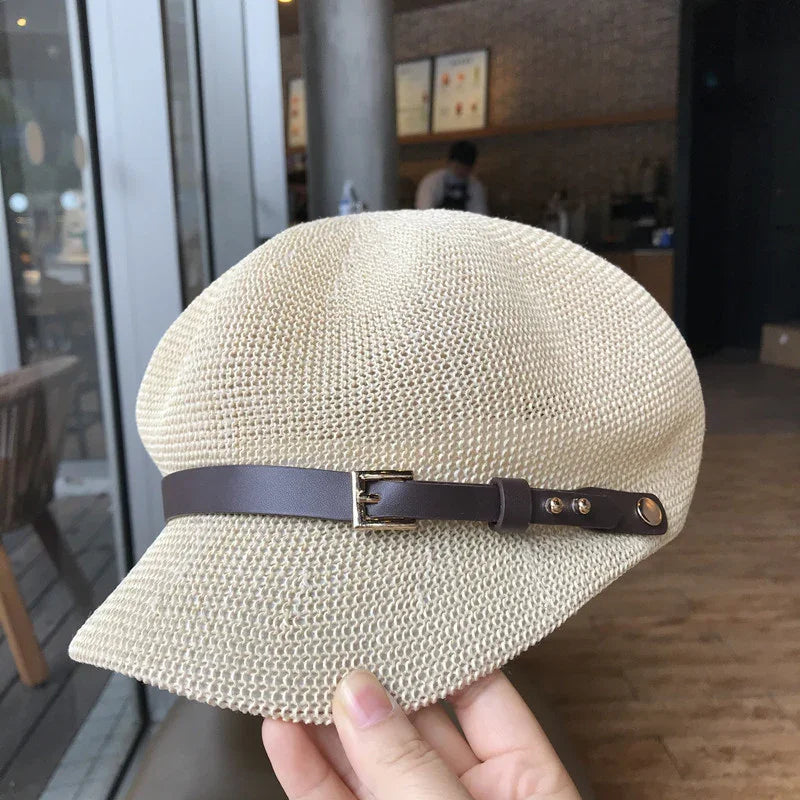 Hand holding beige woven newsboy cap with brown leather strap and gold buckle in cafe interior