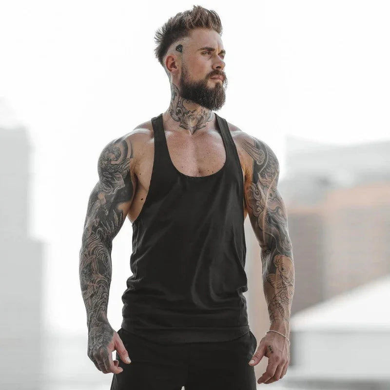 Muscular tattooed man with beard wearing black tank top standing outdoors with blurred city background