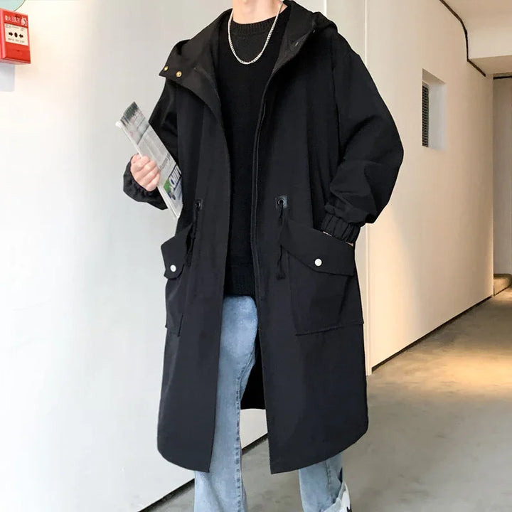 Person wearing black oversized hooded parka coat with flap pockets, light blue jeans, and silver chain necklace indoors