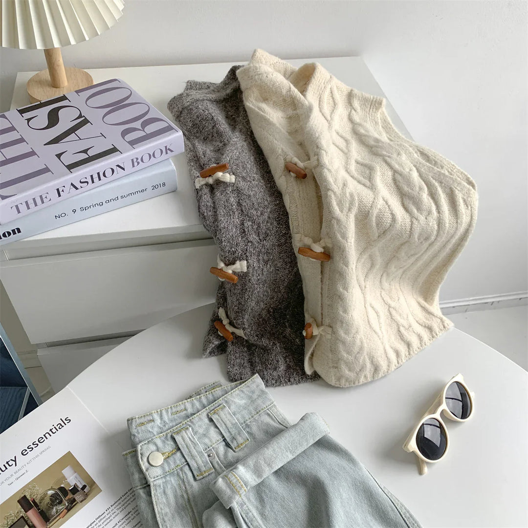 Cozy cable knit vests in cream and gray with toggle buttons on white table with sunglasses, denim, and fashion books