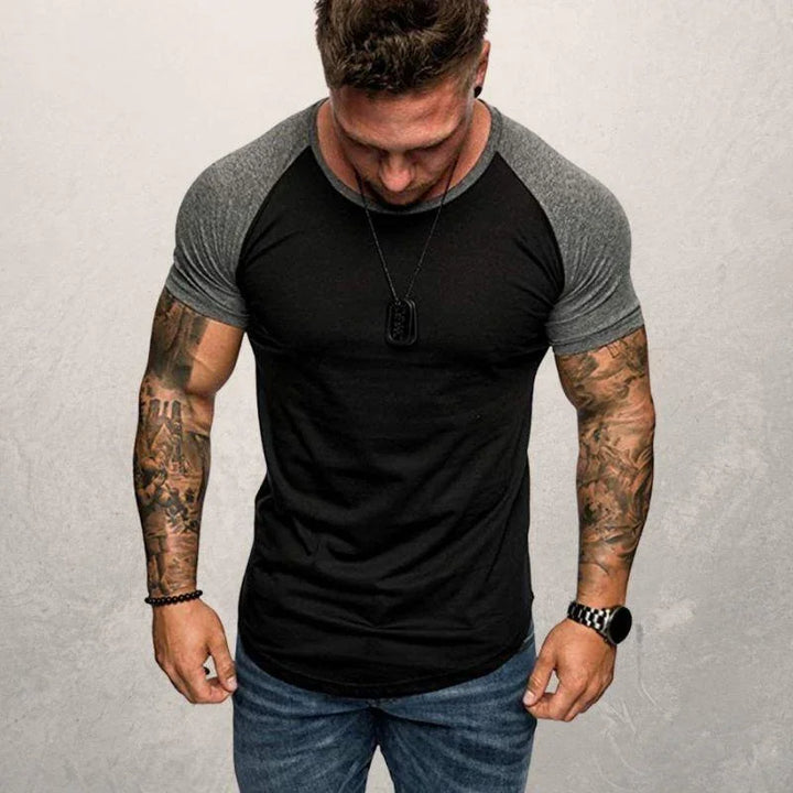 muscular man with tattooed arms wearing black and gray raglan t-shirt and jeans against gray background