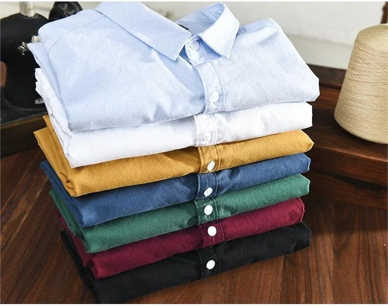Stack of seven folded button-down shirts in light blue, white, mustard, blue, green, burgundy, and black on wooden surface