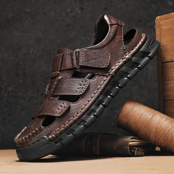 Brown leather men's sandal with black sole and stitched details, displayed with leather craft tools