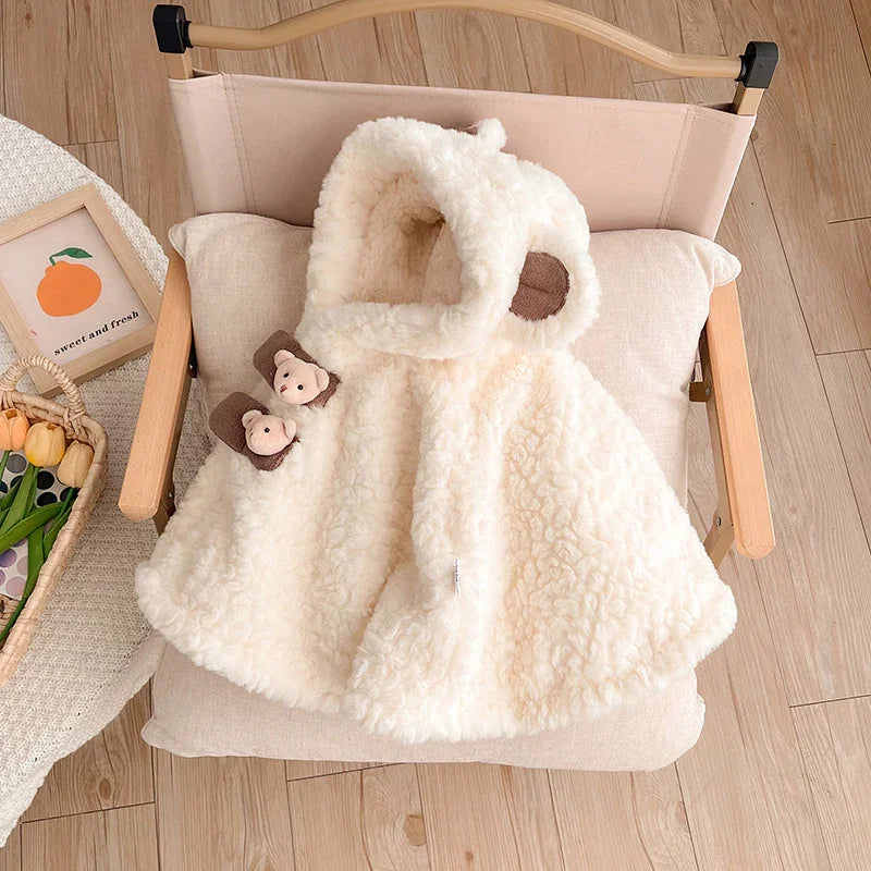 Soft cream-colored fluffy baby hooded cape with bear ears and teddy bear accents on a wooden chair