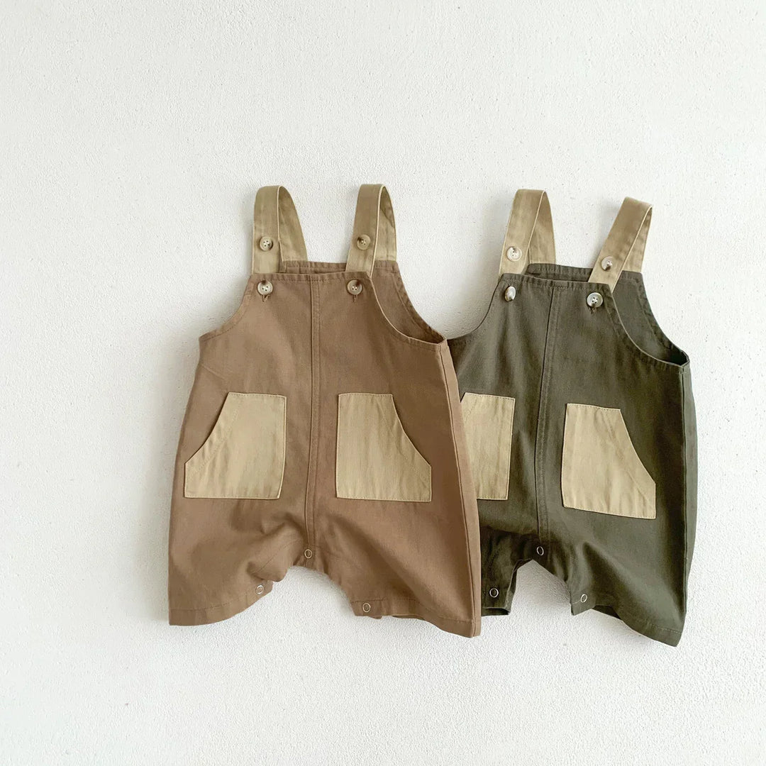 Two baby cotton rompers with beige pockets and straps, in brown and dark green colors, hanging on white wall