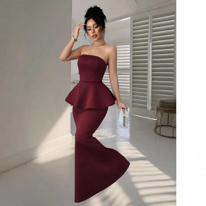 Woman in elegant strapless burgundy peplum mermaid gown holding a silver clutch in modern minimal room