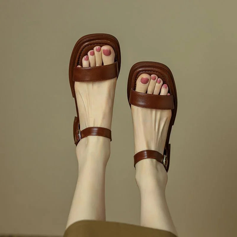 Feet wearing brown leather sandals with square toe design and ankle straps on a beige background