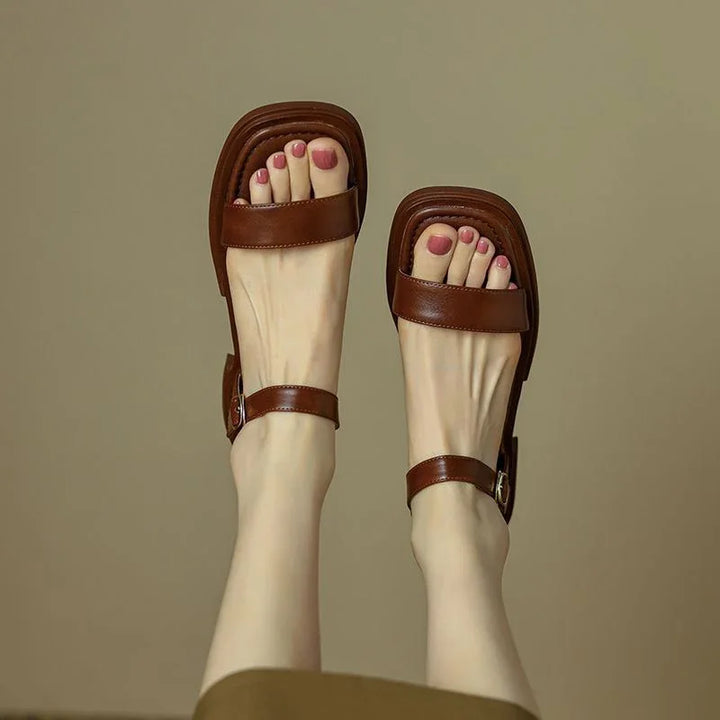 Feet wearing brown leather sandals with square toe design and ankle straps on a beige background
