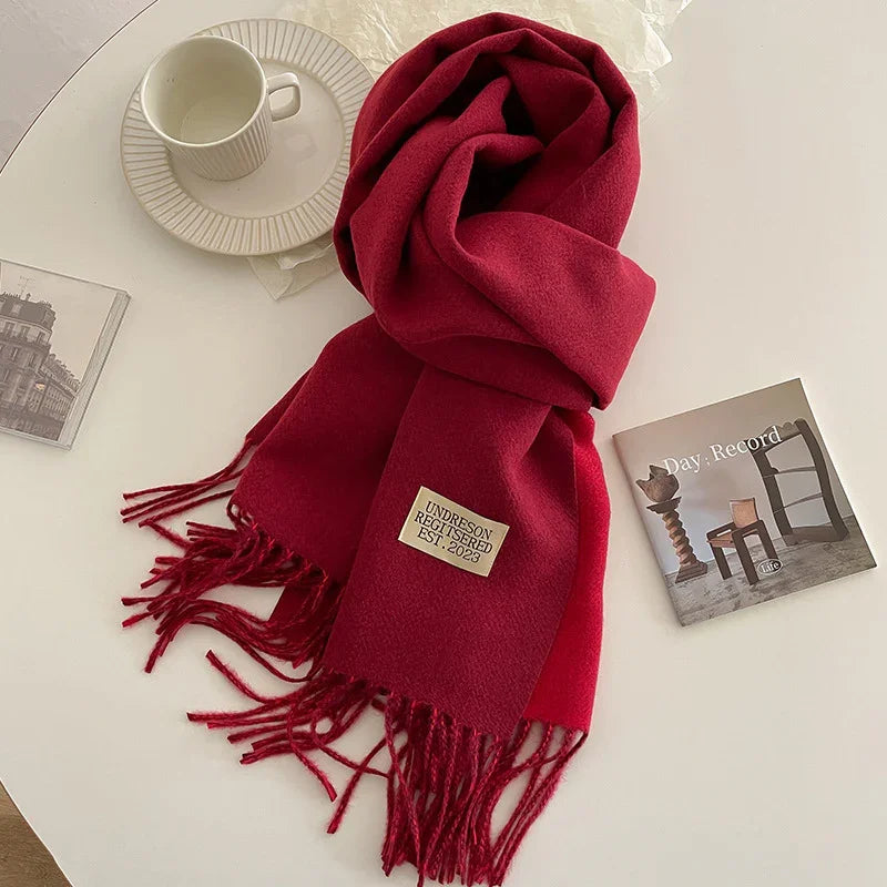 Red woolen scarf with fringe and label 'UNDRESON REGISTERED EST. 2023' on a white table with a cup and magazine