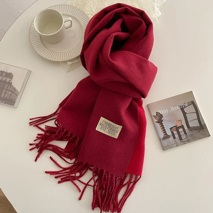 Red woolen scarf with fringe and label 'UNDRESON REGISTERED EST. 2023' on a white table with a cup and magazine