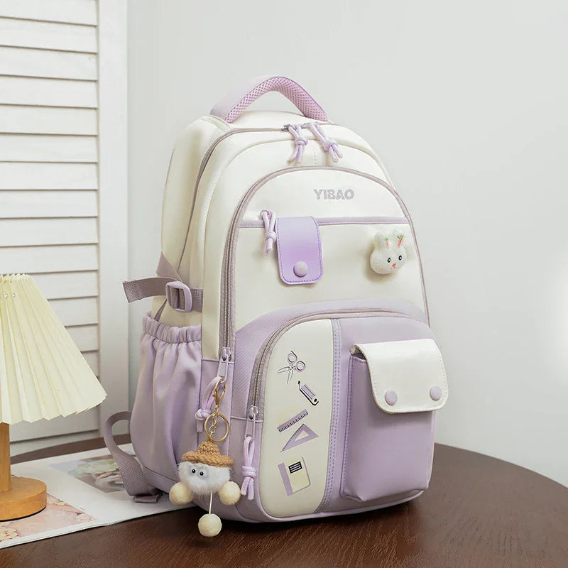Cute white and lavender YIBAO backpack with front pockets, school supplies icons, and plush keychain on wooden table
