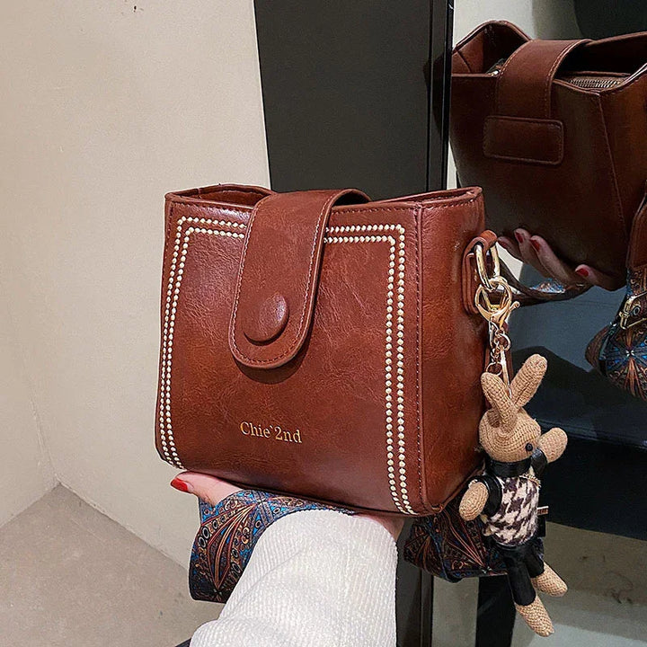 brown leather handbag with studded trim and a knitted bunny keychain held by a hand with red nail polish