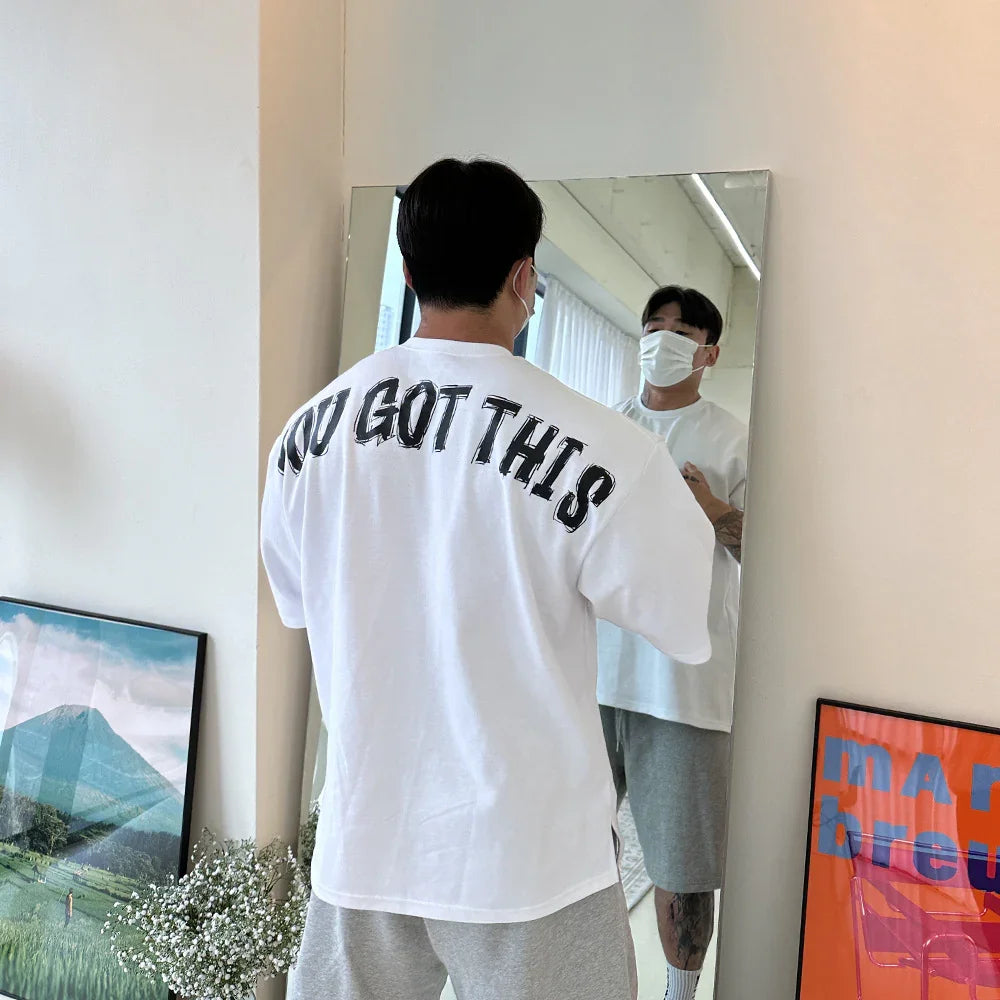 Man wearing white T-shirt with 'YOU GOT THIS' text, gray shorts, standing in front of mirror indoors