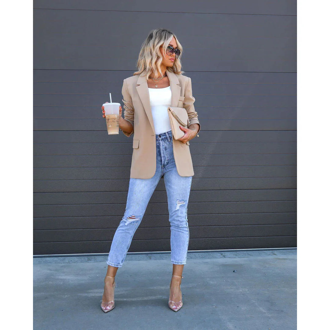 Woman in beige blazer, white top, ripped jeans, clear heels holding coffee and clutch by dark wall