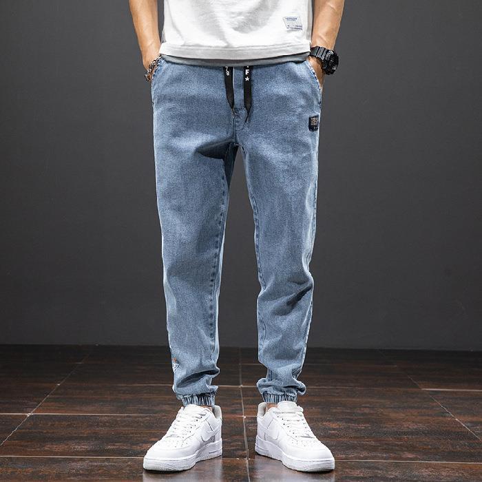 Man wearing light blue denim jogger pants with elastic cuffs and white sneakers, standing on wooden floor