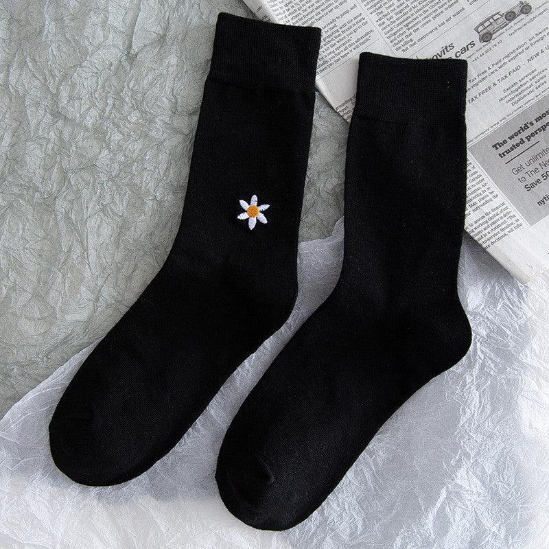 Pair of black socks with small embroidered white daisy flower, lying on crumpled silver fabric and newspaper