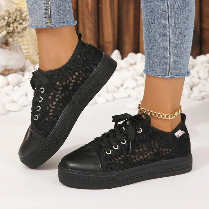 Black lace sneakers with leather toe caps worn with gold anklet and cropped blue jeans on white stone background