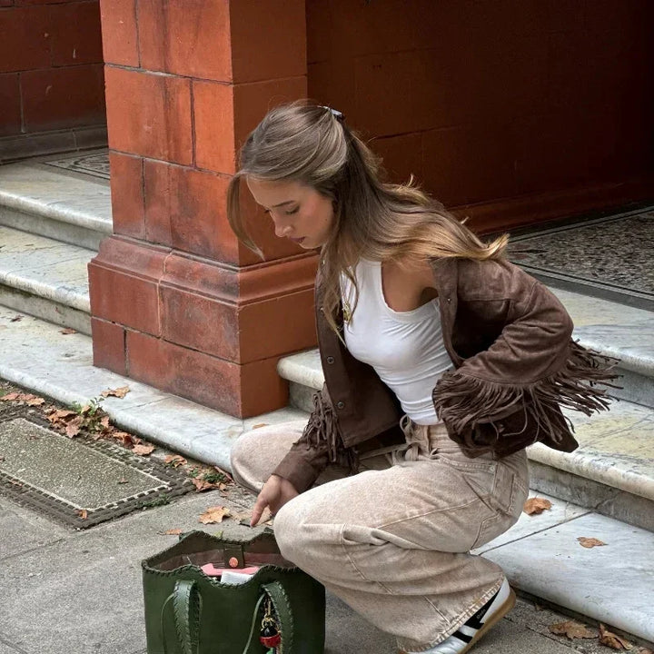 Fashionable woman in beige pants and brown fringe jacket crouching by red brick wall, reaching into large green tote bag