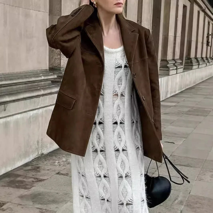 Woman wearing brown suede jacket over white knitted dress holding a black heart-shaped handbag on a stone walkway