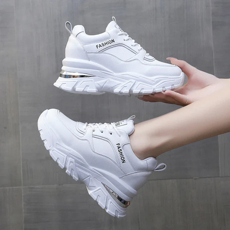 White chunky fashion sneakers with thick soles and metallic accent, worn on feet against gray background