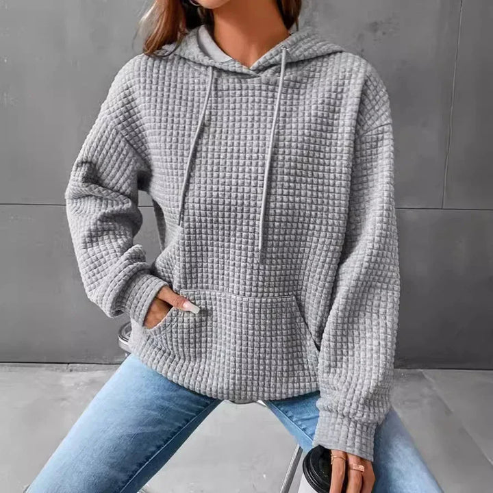 Woman wearing cozy gray waffle knit hoodie with front pocket and light blue jeans sitting on stool