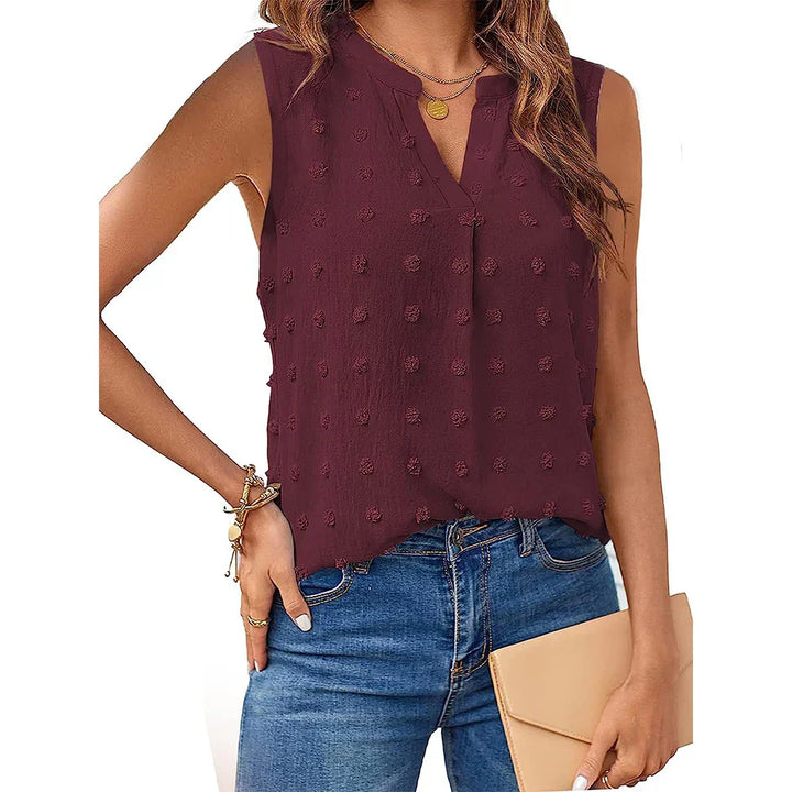 Woman wearing sleeveless burgundy textured blouse with blue jeans and holding beige clutch