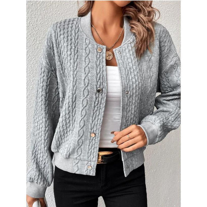 Woman wearing a light gray cable knit bomber jacket with snap buttons, white top, black pants, and gold necklace
