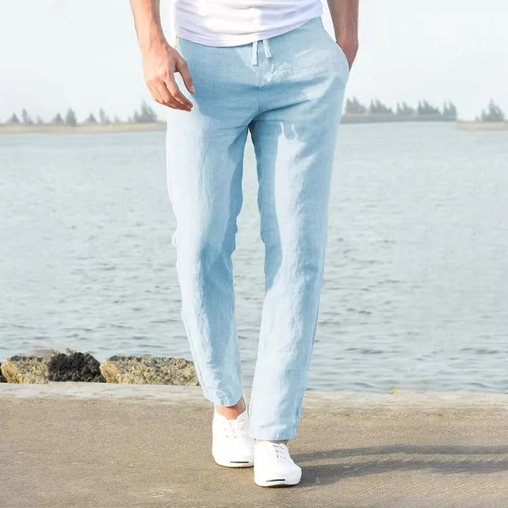 Man wearing light blue linen pants and white sneakers walking by waterfront on clear day