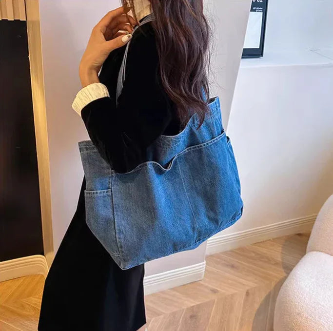 Woman wearing black outfit carrying large blue denim tote bag indoors on wooden floor