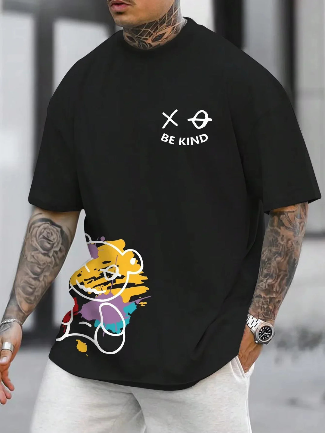 Man wearing black t-shirt with white BE KIND text and multicolor bear graphic, tattoos on arms