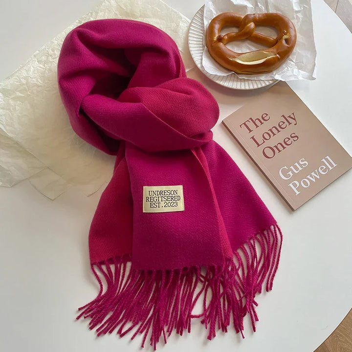 Bright pink fringed scarf with Undreson Registerered label beside book The Lonely Ones by Gus Powell and a soft pretzel on white table