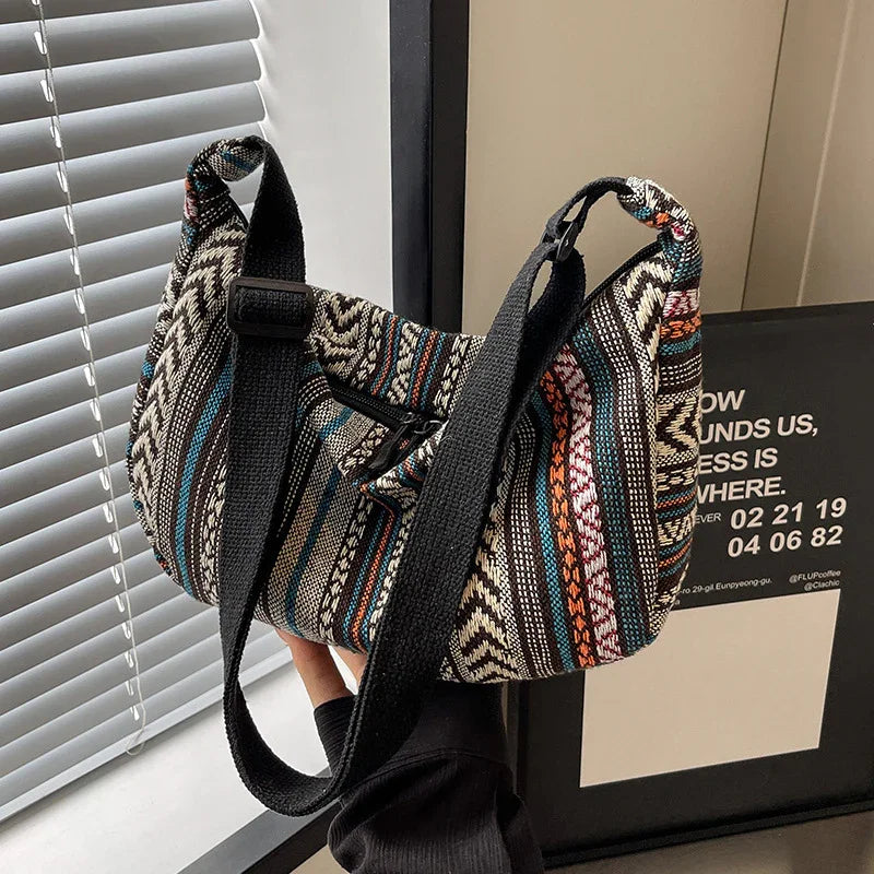 Hand holding colorful woven shoulder bag with black adjustable strap near window blinds and framed wall art