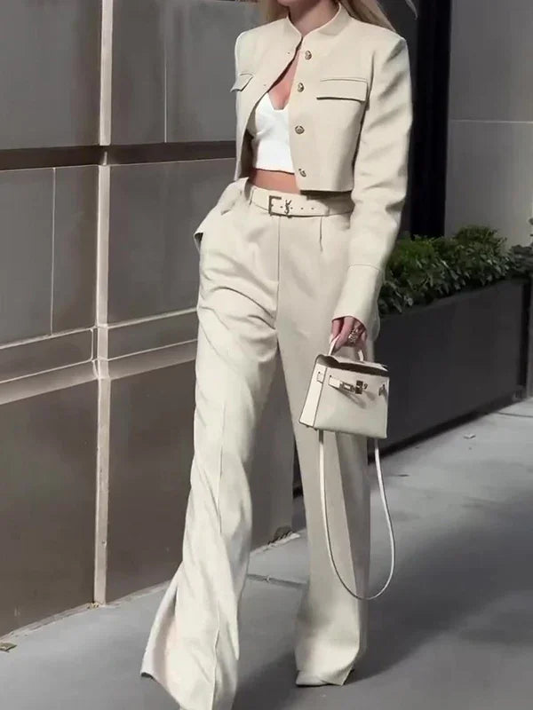 Woman in beige cropped blazer and high-waisted trousers carrying a small beige handbag on city street