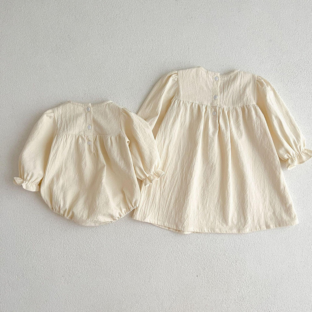 Two cream-colored baby outfits including a long-sleeve dress and a matching romper with buttons on light background