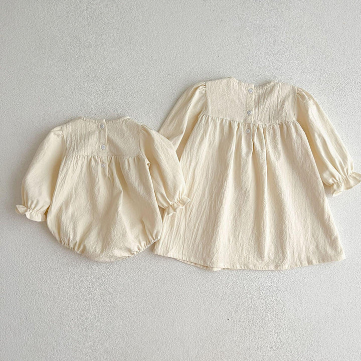 Two cream-colored baby outfits including a long-sleeve dress and a matching romper with buttons on light background