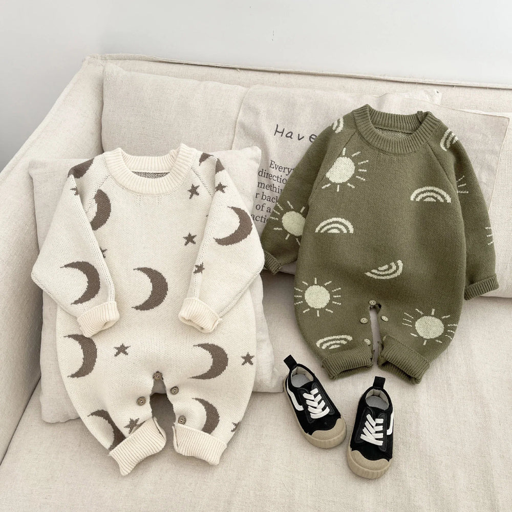 Two knitted baby rompers on beige couch, one cream with moons and stars, one green with suns and rainbows, black baby sneakers