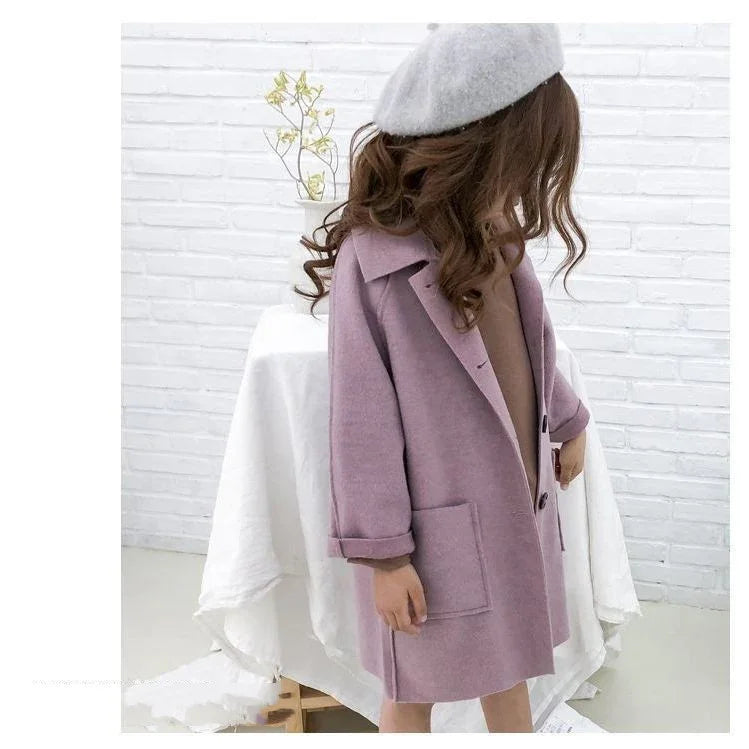 Child wearing a lavender wool coat and gray beret standing indoors against a white brick wall