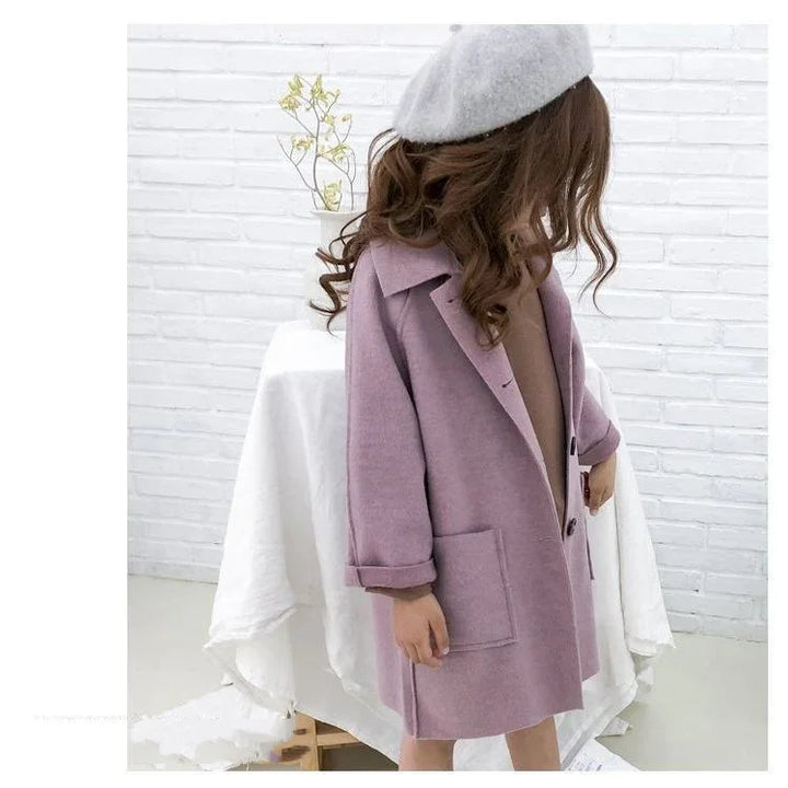 Child wearing a lavender wool coat and gray beret standing indoors against a white brick wall