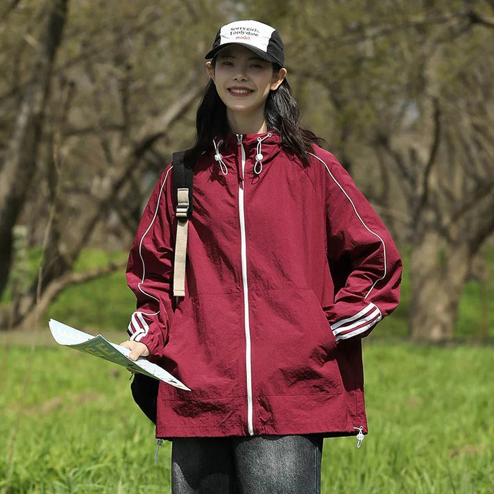 Smiling woman in maroon windbreaker jacket and black cap holding map in outdoor green park
