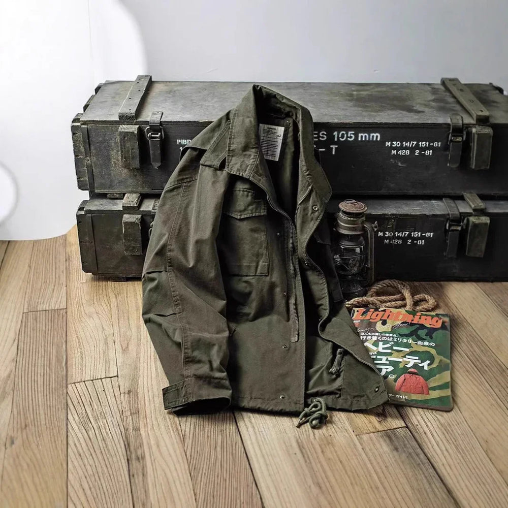 Green military jacket displayed on wooden floor with vintage metal ammo boxes, lantern, and magazine