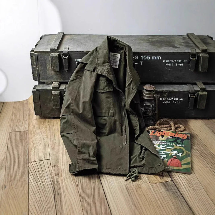 Green military jacket displayed on wooden floor with vintage metal ammo boxes, lantern, and magazine