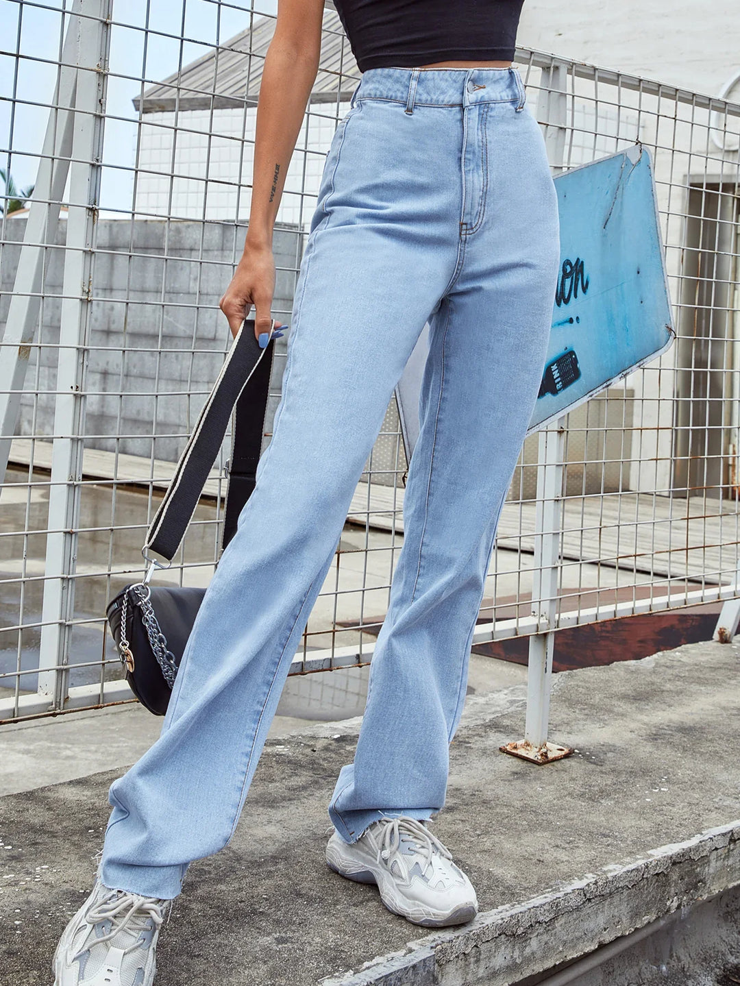 Close-up of person wearing light blue high-waisted jeans, white sneakers, black crop top, and holding black handbag