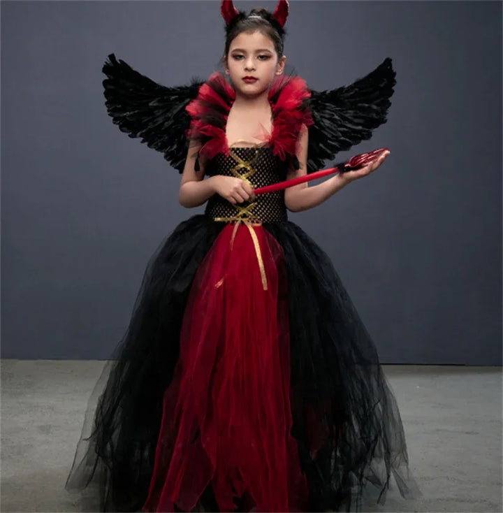 Young girl in black and red devil costume with wings and trident standing against gray background