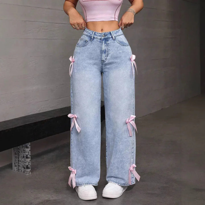 Front view of light blue wide-leg jeans with pink ribbon bows worn with white sneakers indoors
