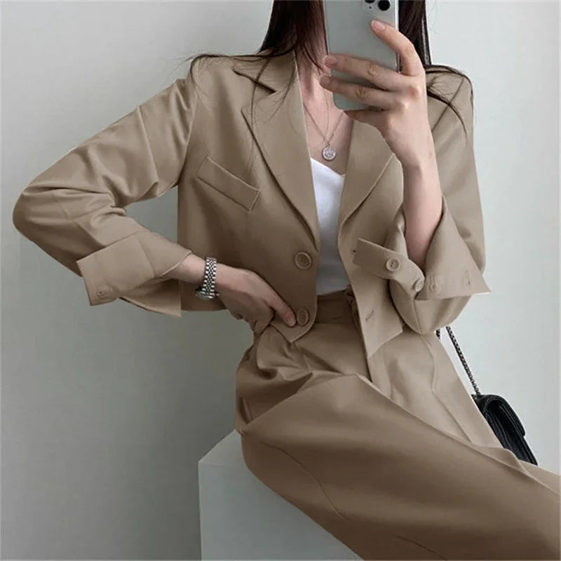 Woman wearing beige cropped blazer and matching high-waisted trousers, holding phone for mirror selfie