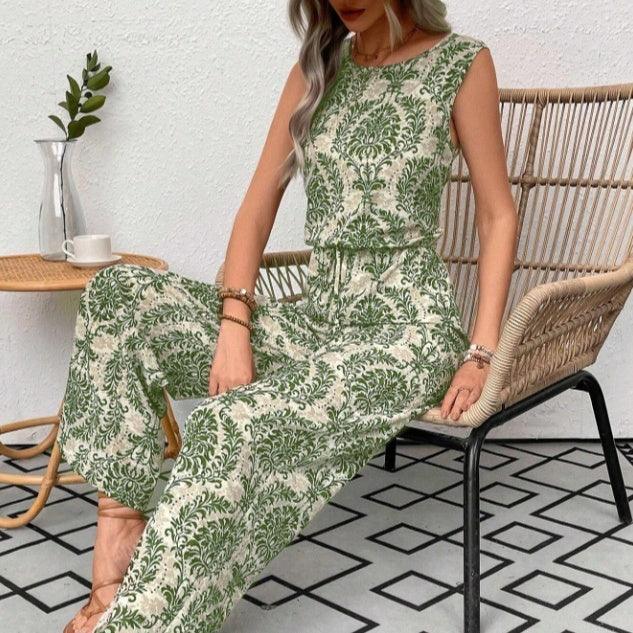Woman wearing green and white sleeveless floral jumpsuit sitting on woven chair beside small wooden table with glass vase